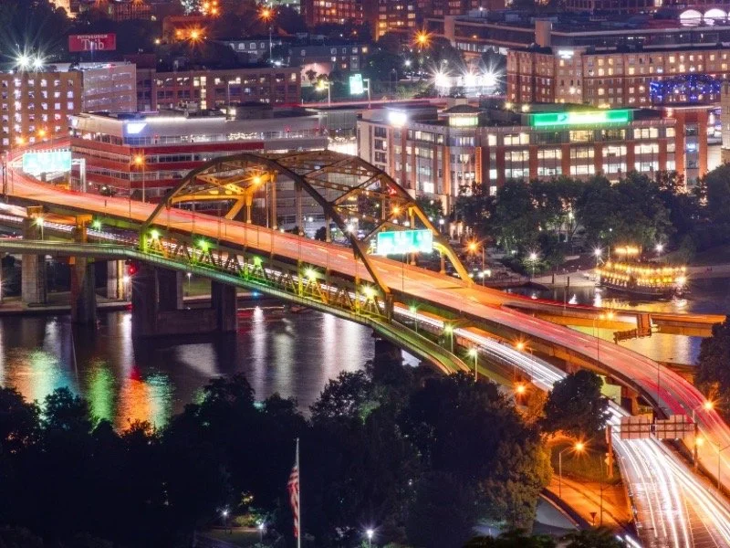 Night view of Pittsburgh's downtown skylone with the Fort Pitt Bridge illuminated, reflecting the city's rivers, bridges, and vibrant urban energy after dark