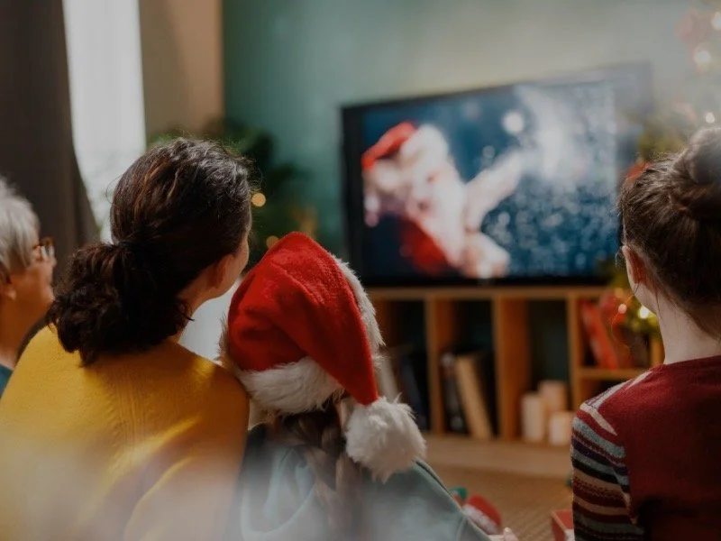 A family sitting on a couch watching a Christmas movie on TV, with warm lighting, festive decor, and a child wearing a Santa hat, capturing a cozy holiday movie night atmosphere.