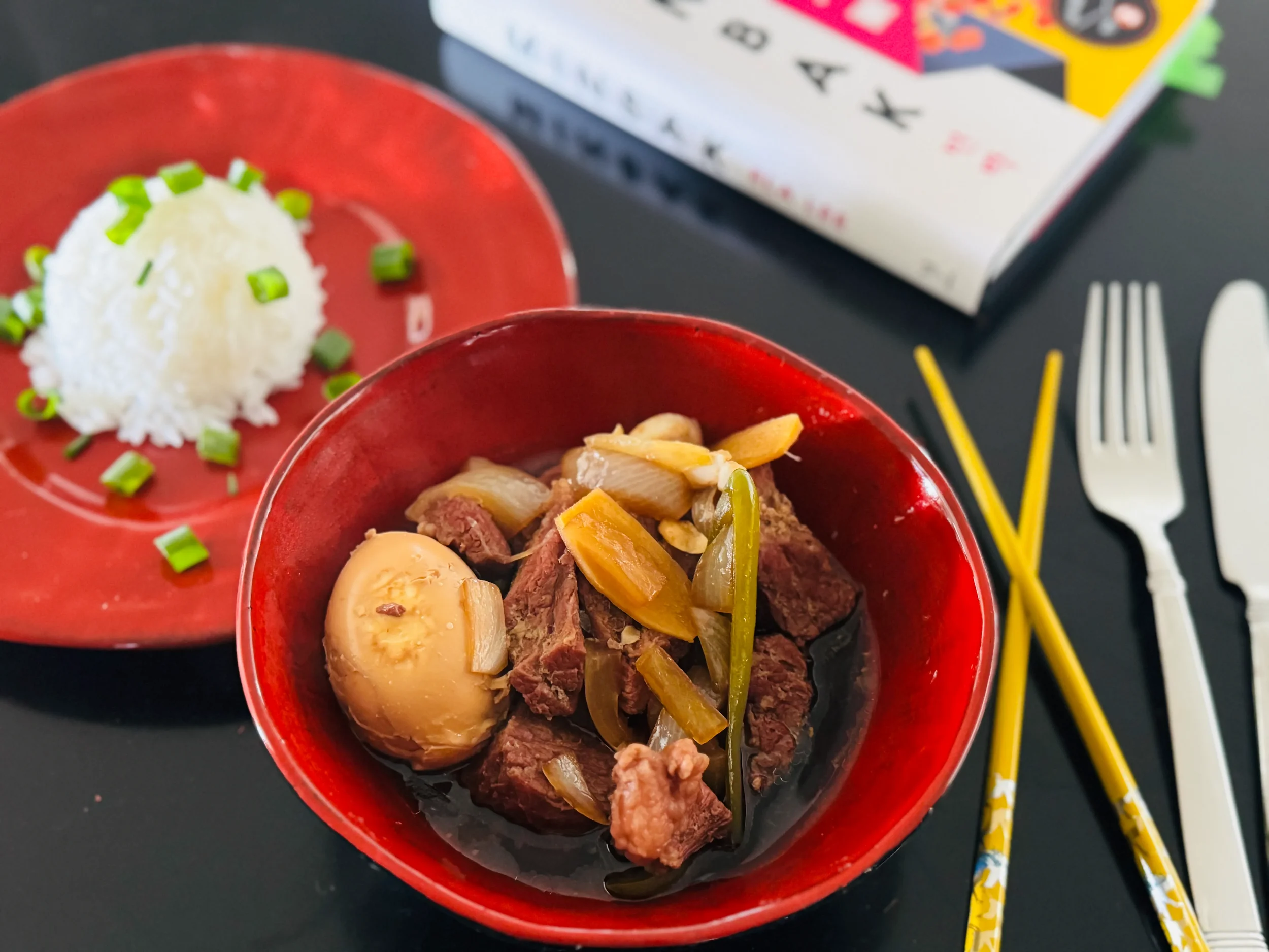 A red bowl of Korean jangjorim braised beef with soy-stained eggs and vegetables, served alongside steamed rice with scallions, chopsticks, fork, and the novel Minbak in the background