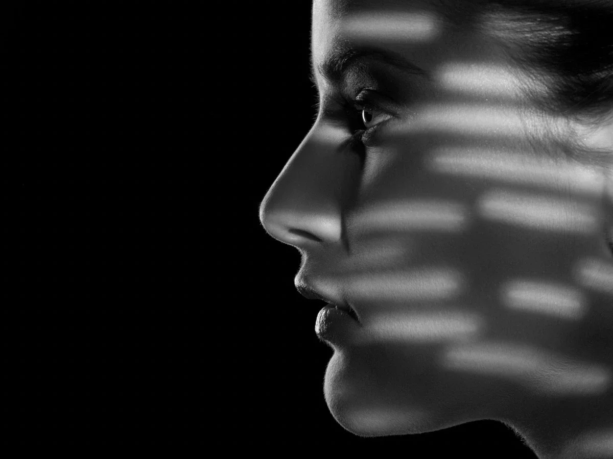 Black and white close-up profile portrait of a woman's face partly obscured by striped shadow, representing being seen but not fully known
