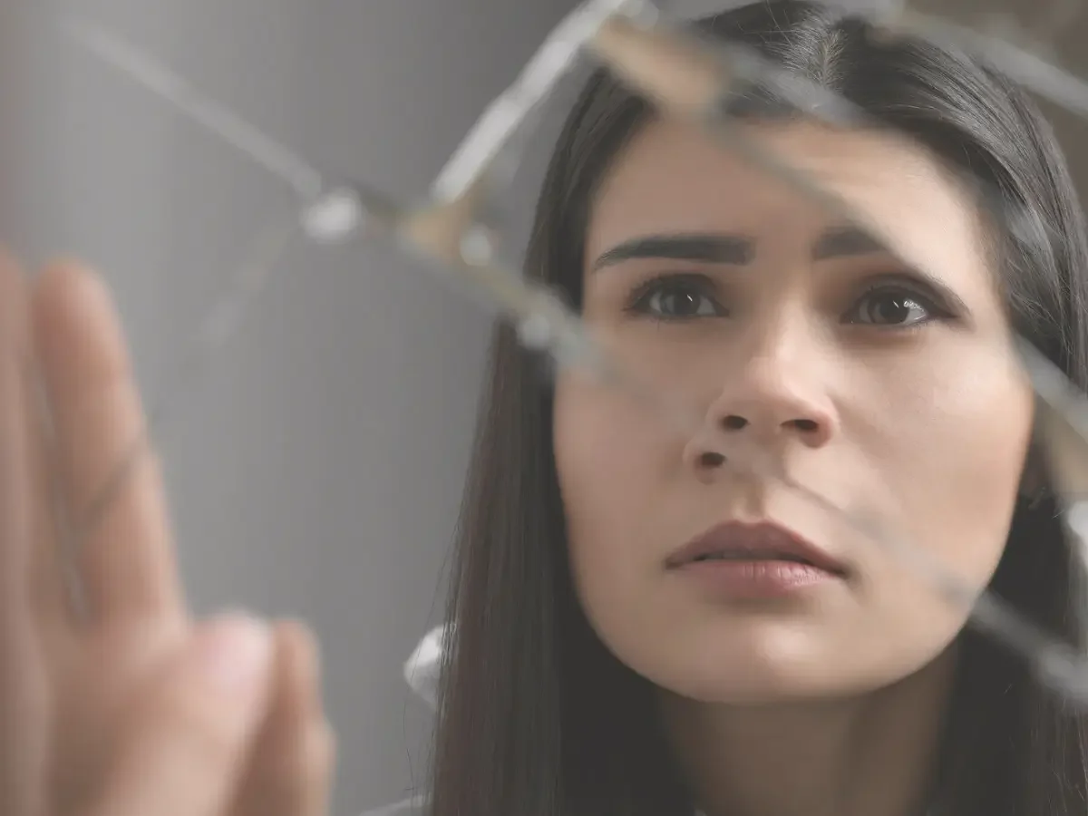 A woman's face seen through a shattered mirror, her hand touching the fractured glass, gaze steady rather than distressed. Accompanying "When the Villain Reflects Us Back" editorial essay.