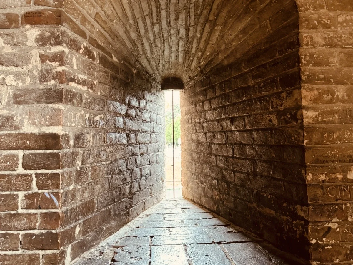 A narrow stone corridor inside Castello Estense, the moated Renaissance castle in Ferrara, Italy, with a single sliver of daylight visible at the far end — the real seat of the Este court Lucrezia de' Medici entered as Alfonso's bride.