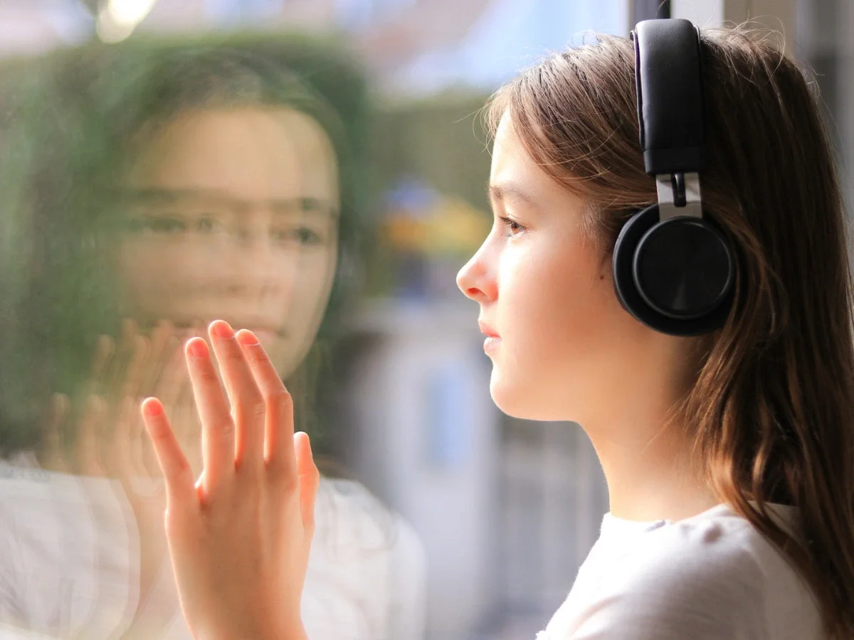 Young girl wearing headphones gazes out a window, her reflection visible in the glass, evoking quiet emotional reflection while listening to music