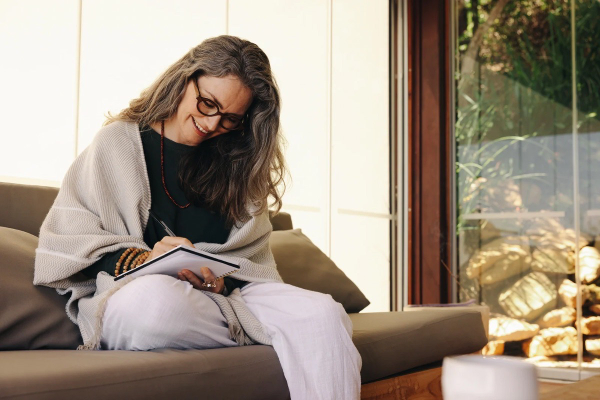 A woman sitting comfortably indoors writing in a journal, representing mindful reflection, stress awareness, and intentional living practices that promote clarity and emotional balance.
