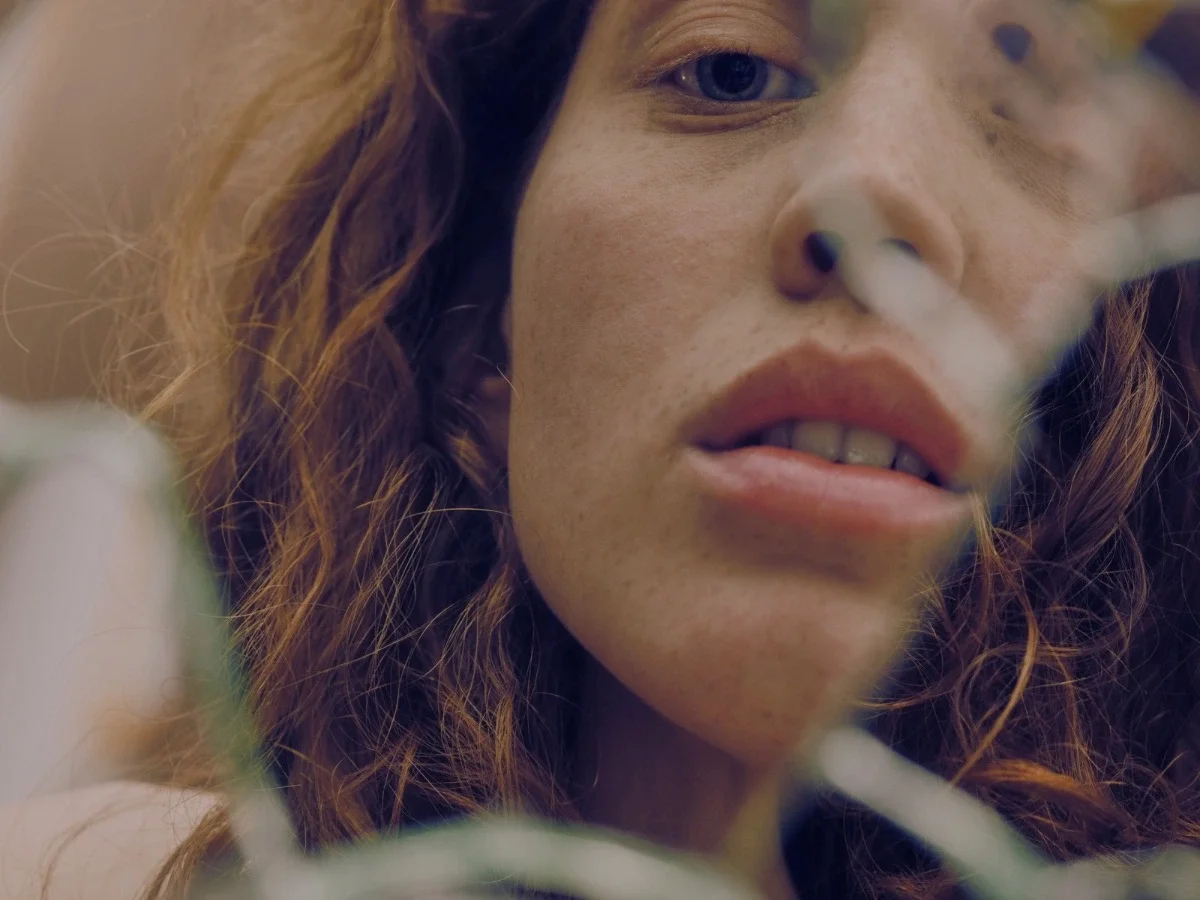 Close-up of a woman's face partially obscured by branches, mouth slightly open as if speaking — illustrating the experience of being expressive and still not fully seen.