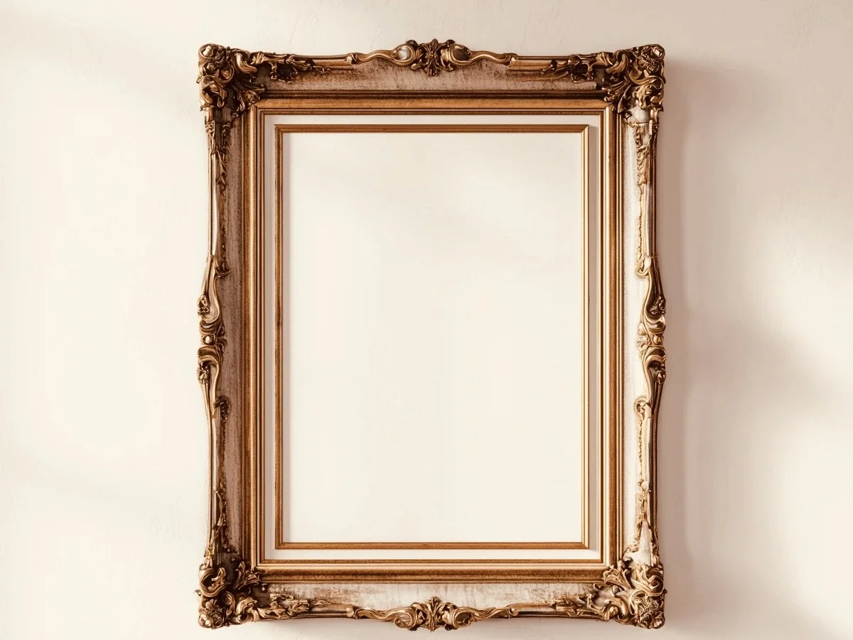 An elaborate gilt frame hangs empty on a warm off-white wall, its ornate carved detail in sharp contrast to the blank space within it. Natural light falls across the wall. Nothing has been chosen to fill it.