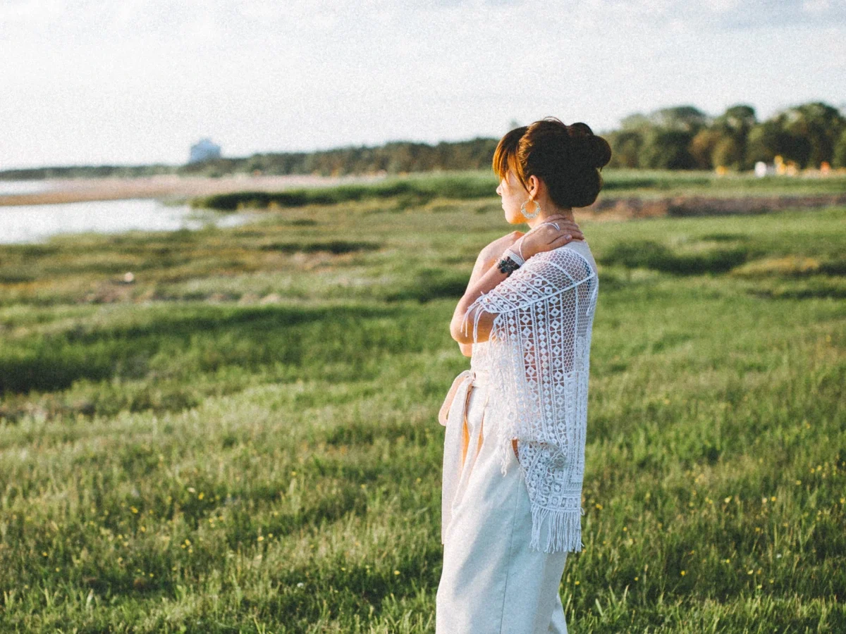 A woman standing alone in an open field, arms wrapped around herself, facing the horizon