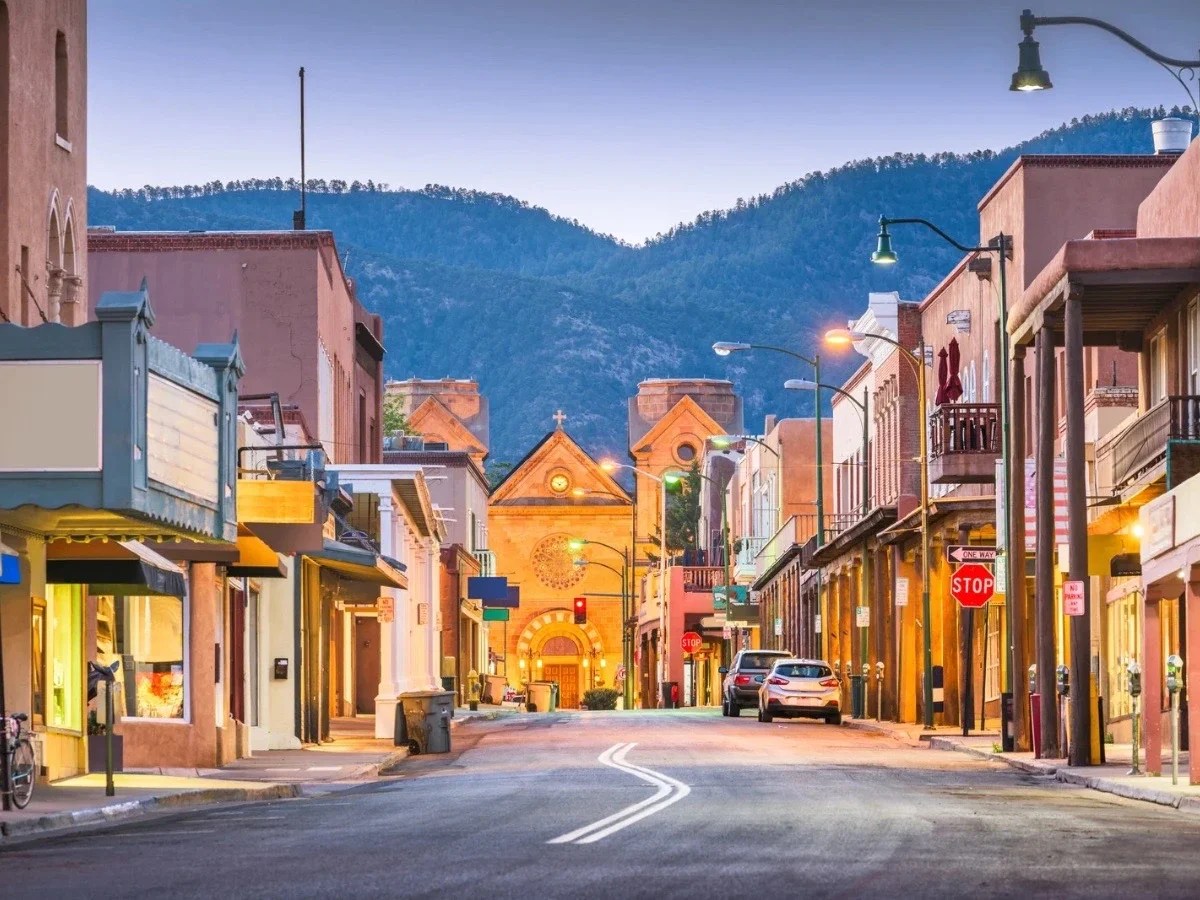 Santa Fe's historic adobe-lined streets at dusk with the Cathedral Basilica and Sangre de Cristo Mountains in the background.