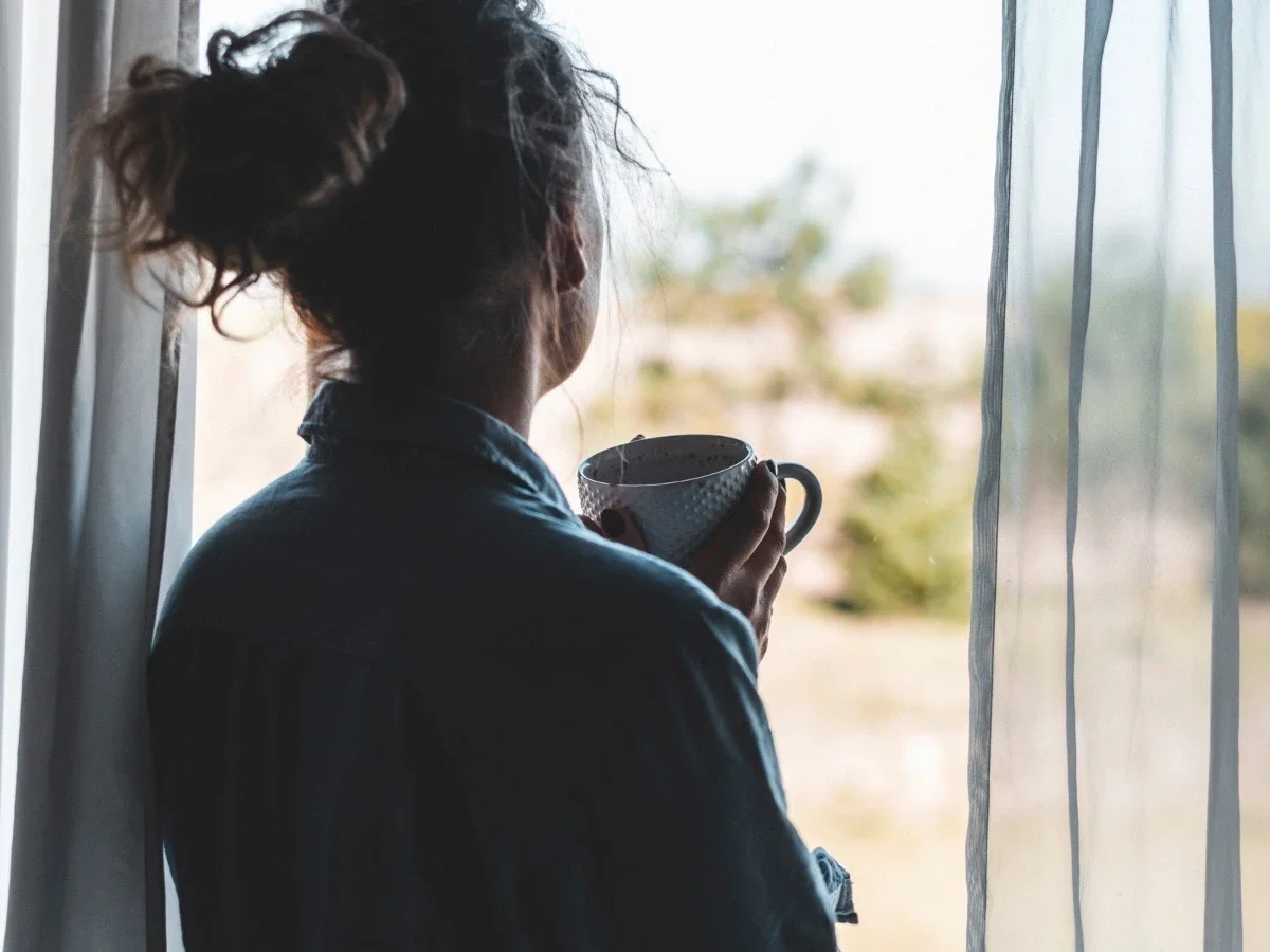 A woman stands at a sunlit window in a darkened room, seen from behind, holding a warm mug with both hands. She gazes outside without drinking, caught in a moment of quiet interior suspension.