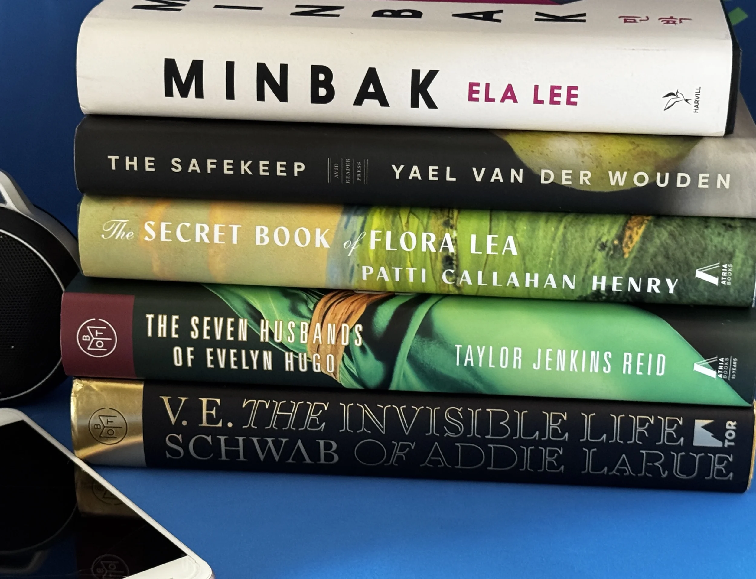 Stack of five novels including Minbak, The Safekeep, and Addie LaRue alongside a JBL speaker and phone — literary playlist pairings