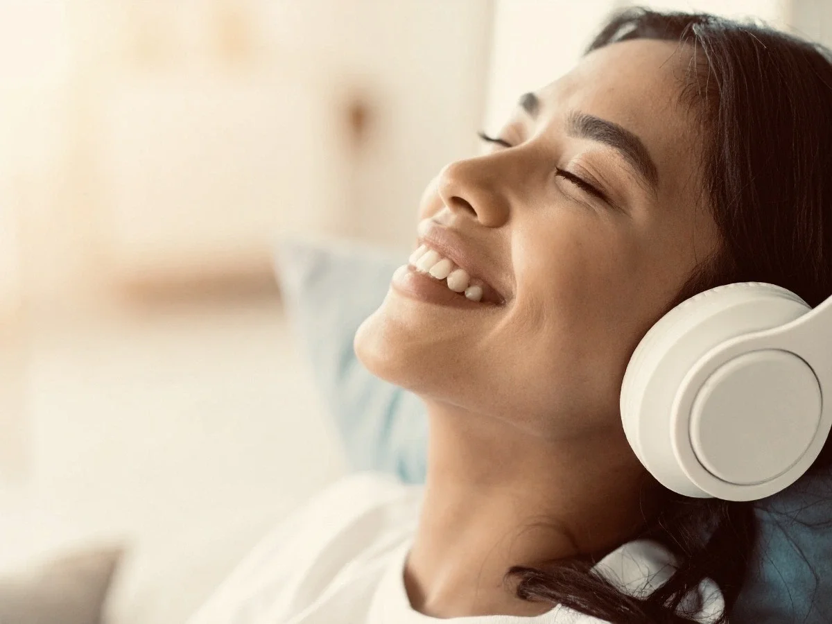 A woman wearing white over-ear headphones reclines with her eyes closed and a relaxed smile, bathed in soft warm light