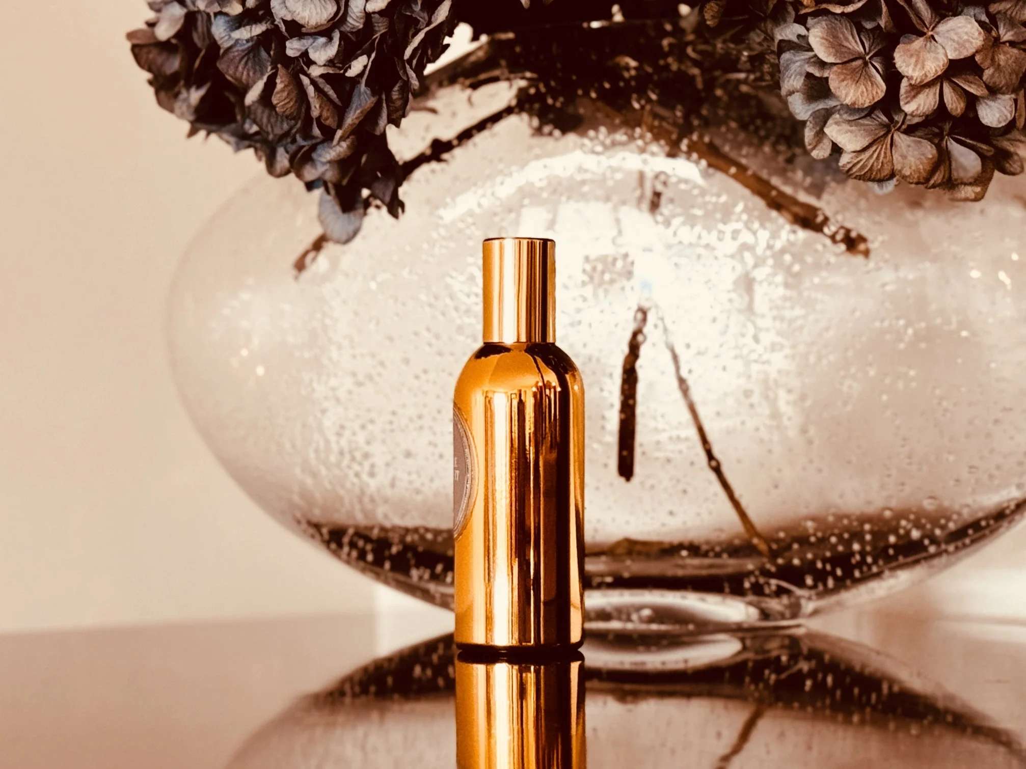 Single gold perfume bottle on a reflective surface with dried blue hydrangeas, representing the intentional process of choosing a signature scent