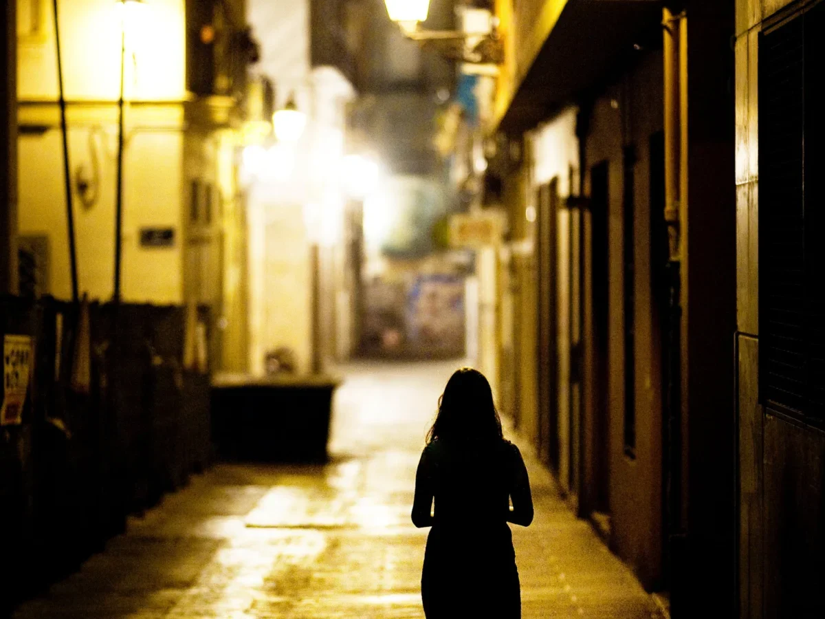 Silhouette of a woman walking alone down a narrow fog-lit urban street at night, seen from behind