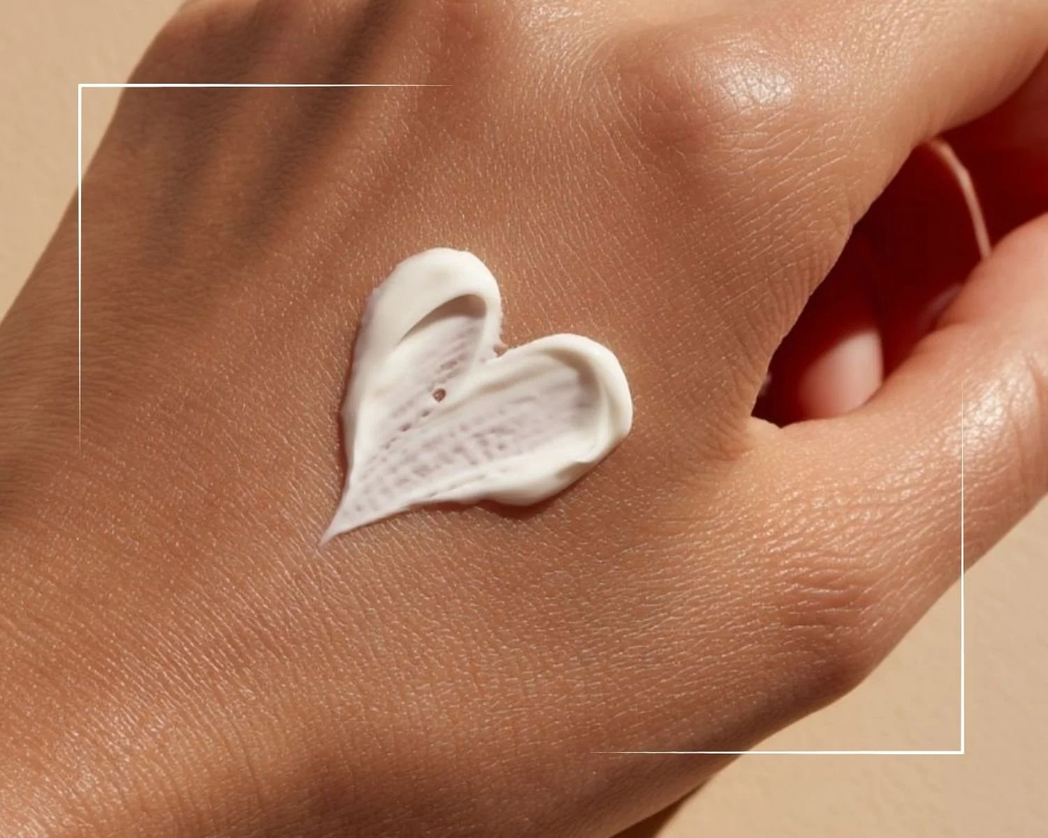 A close-up of a hand with smooth skin, with a small amount of white cream shaped like a heart applied on the back of the hand against a neutral beige background.