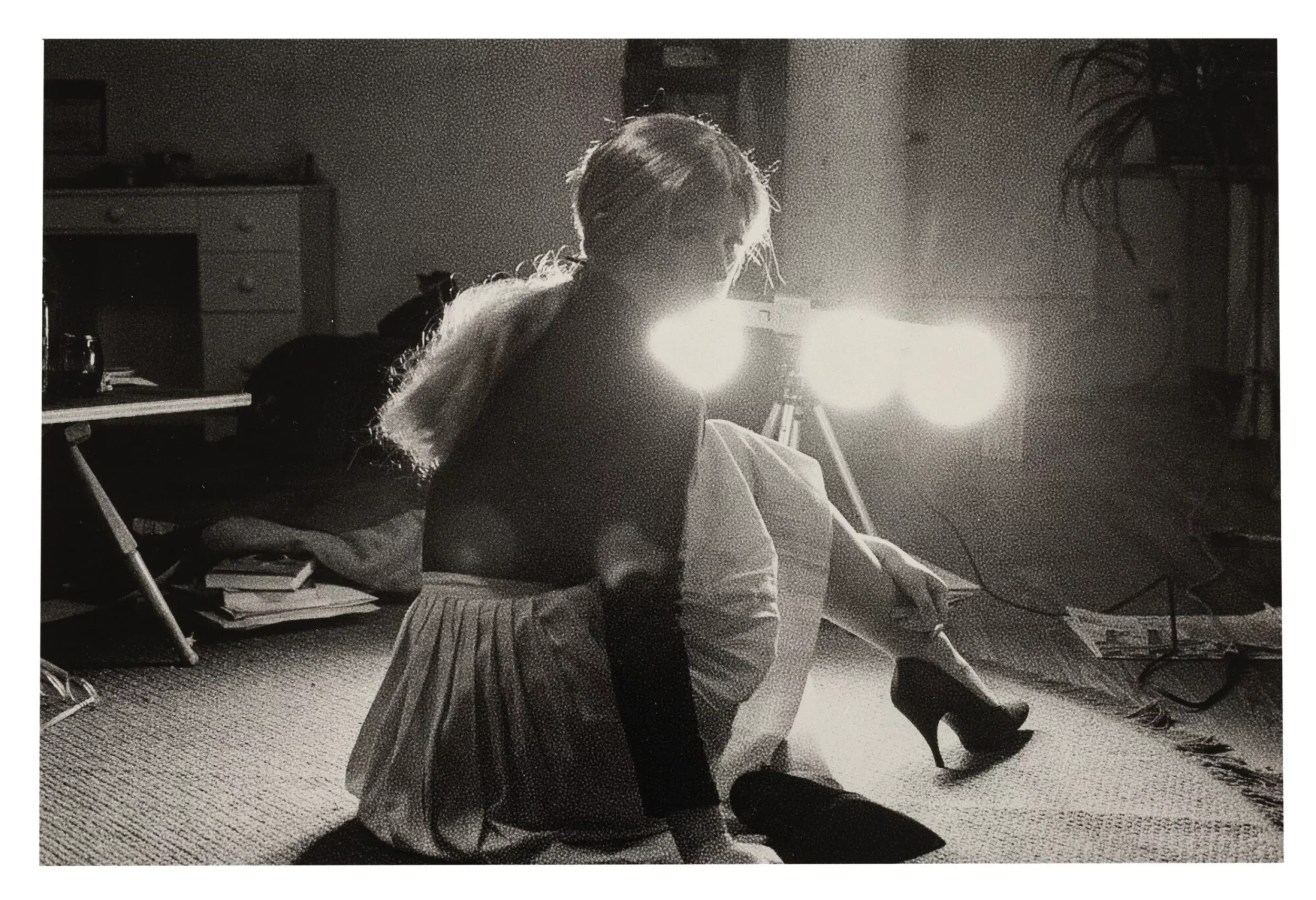 Black-and-white photograph by Cindy Sherman showing a woman seated on the floor in high heels facing bright studio lights, examining performance, constructed identity, and cinematic self-image.
