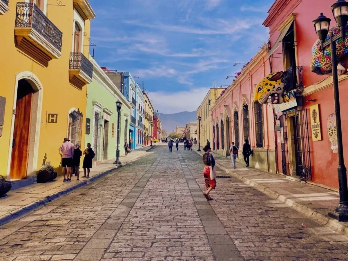 Pedestrians walking along a colorful cobblestone street in Oaxaca's historic center, lined with colonial buildings and local shops.