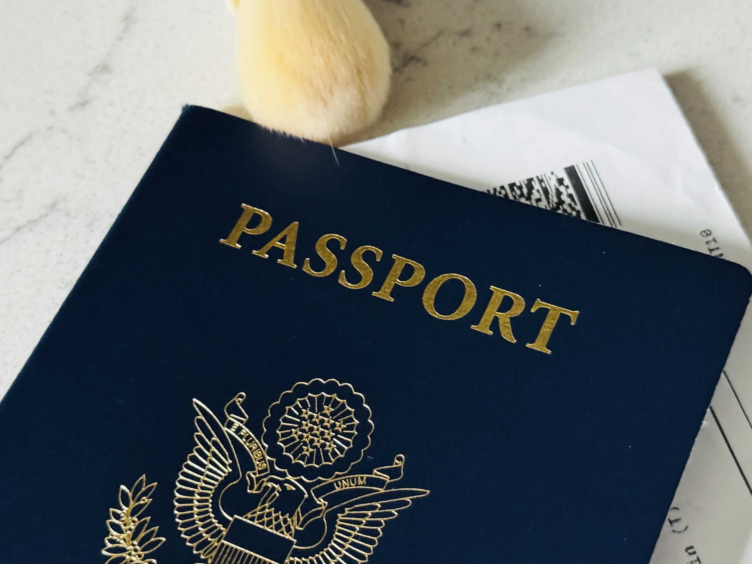 Passport and makeup brush on marble surface representing travel skincare packing essentials