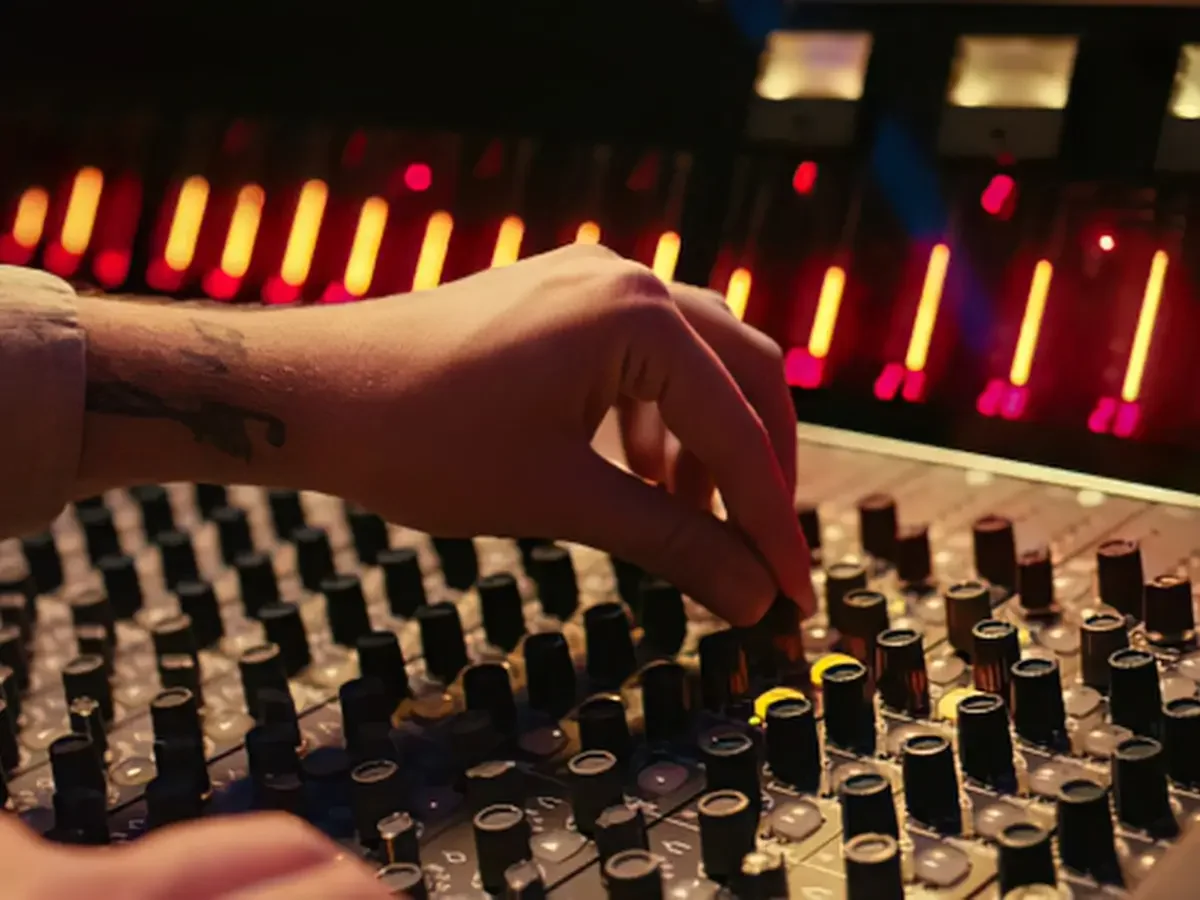A hand on one fader of a studio mixing console, with the rest of the board softly blurred.