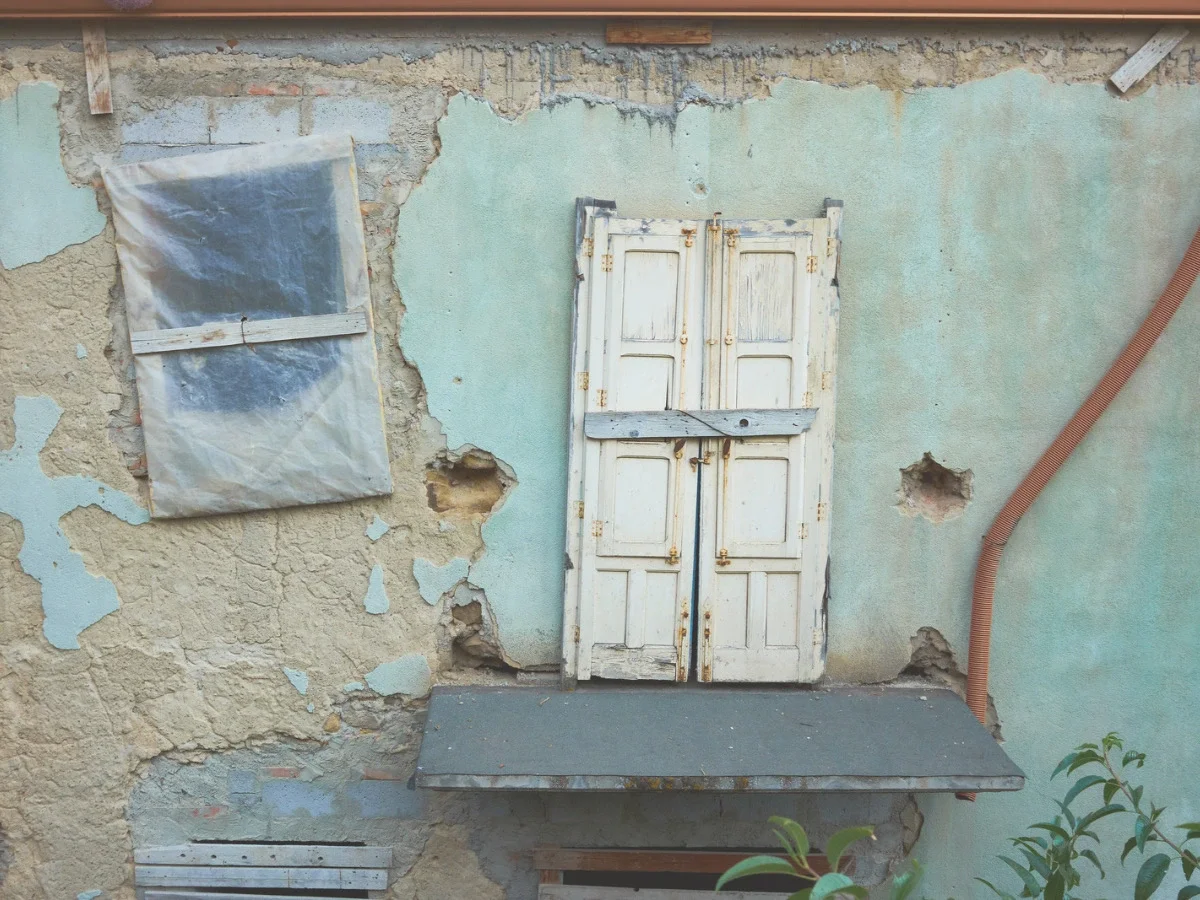 A weathered exterior wall with peeling paint and a closed wooden shutter, photographed with visible age and imperfection — closing image for She The King's essay "The Desire We Keep Calling Impractical."