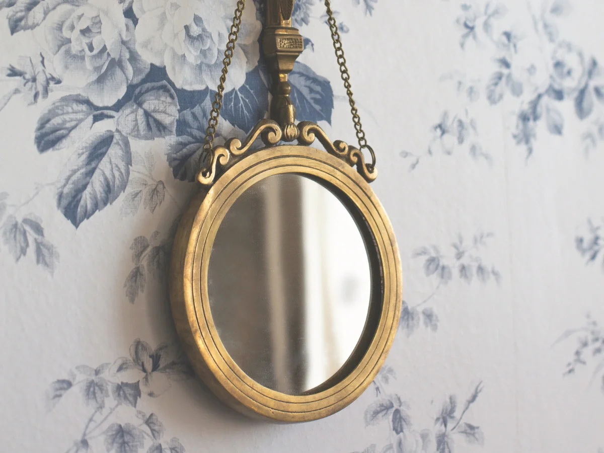 A small gilt oval mirror hanging by chains against vintage blue floral wallpaper, reflecting a softly blurred interior — opening and closing image for She The King's essay "What Iris Apfel Knew About Getting Dressed."