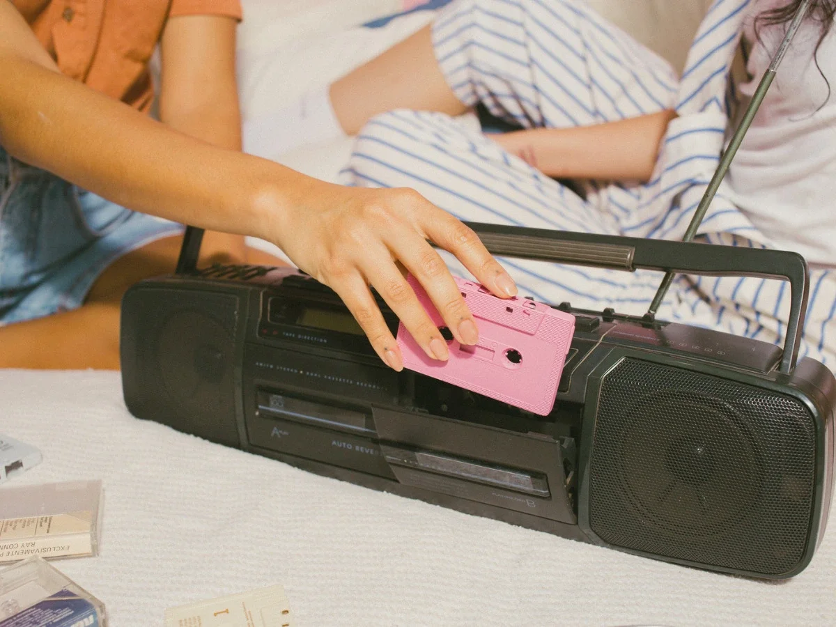 Hand loading a pink cassette tape into a boombox while sitting close to another person, evoking the songs permanently linked to a specific relationship or memory