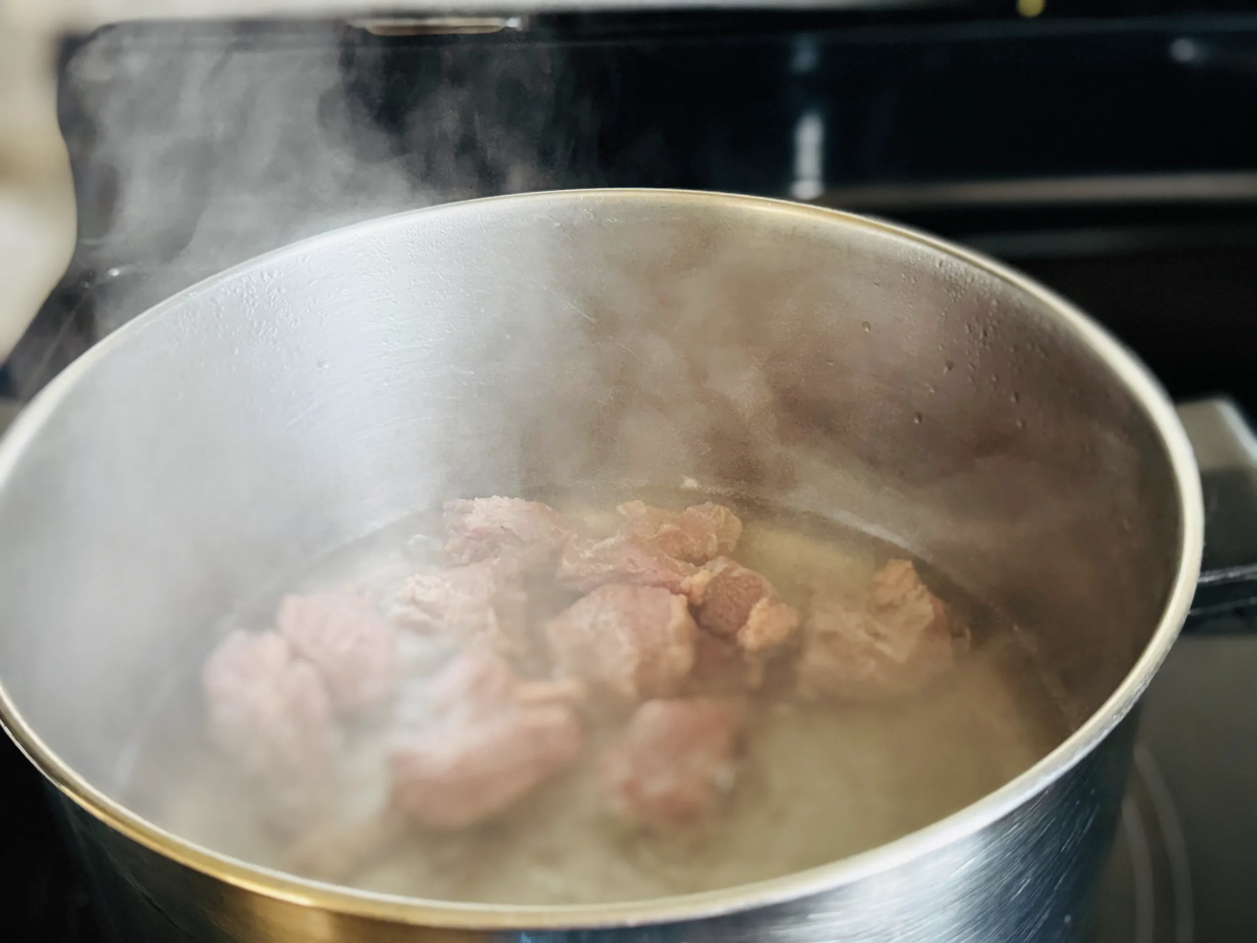 Beef brisket pieces blanching in a stainless steel pot of steaming water on a stovetop, the first step in making Korean jangjorim braised soy sauce beef