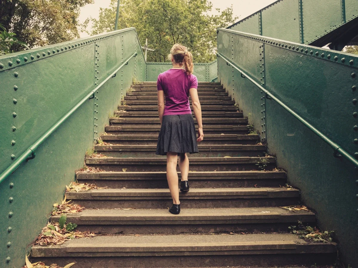 Woman in a purple top and skirt climbing a long outdoor staircase alone, seen from behind, symbolizing self-reliance and emotional distance