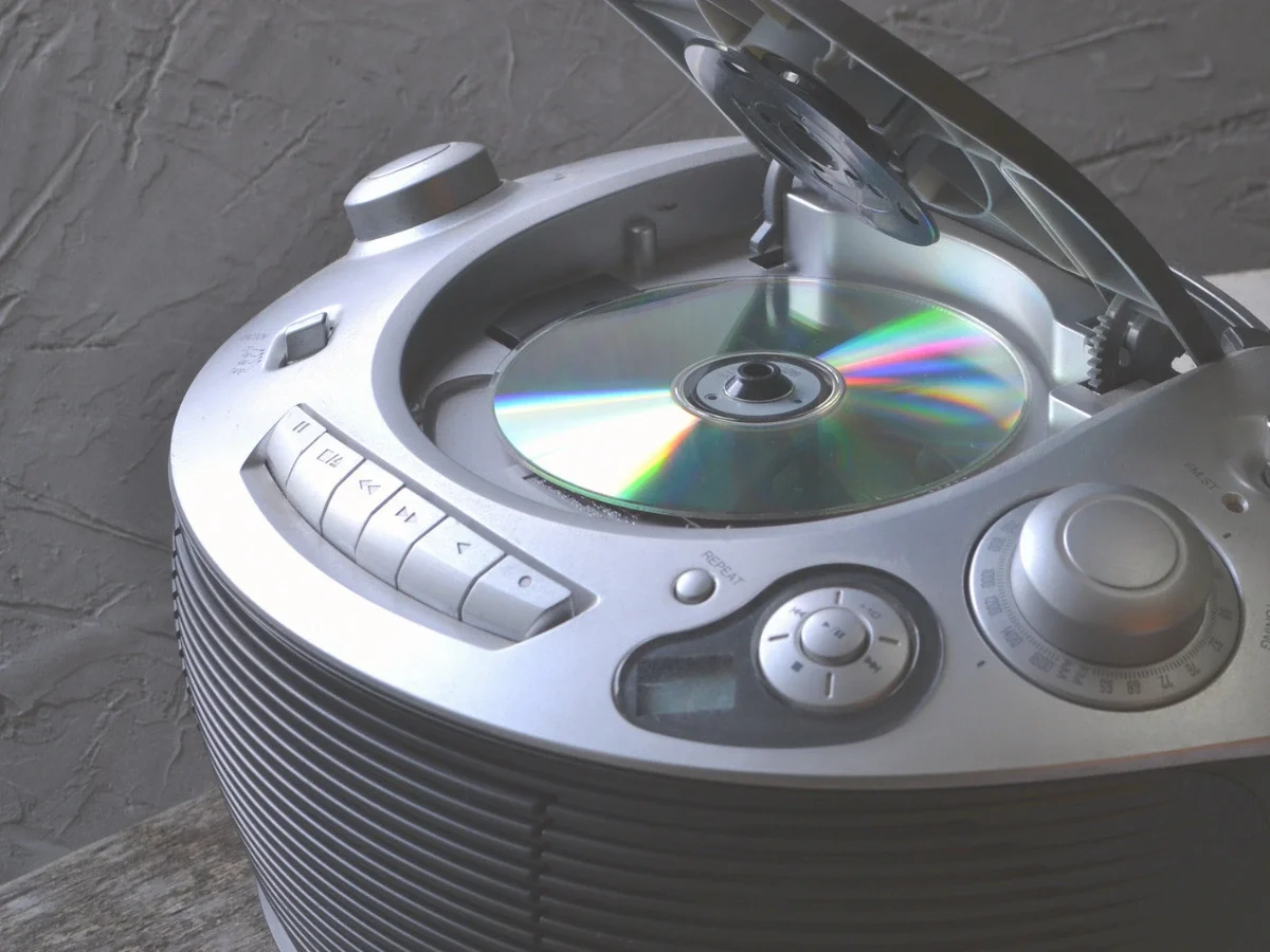 A worn silver portable CD player with the lid open, revealing a reflective disc catching rainbow light—accompanying "The CD Makes It Cleaner," the section of She The King's essay on the shift from vinyl to digital formats.