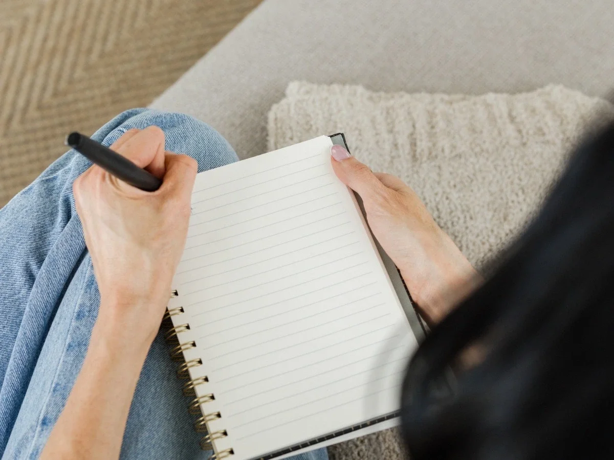 Close-up of a woman's hands holding an open blank notebook with a pen, seated on a couch — representing the quiet, private act of examining your inner voice.