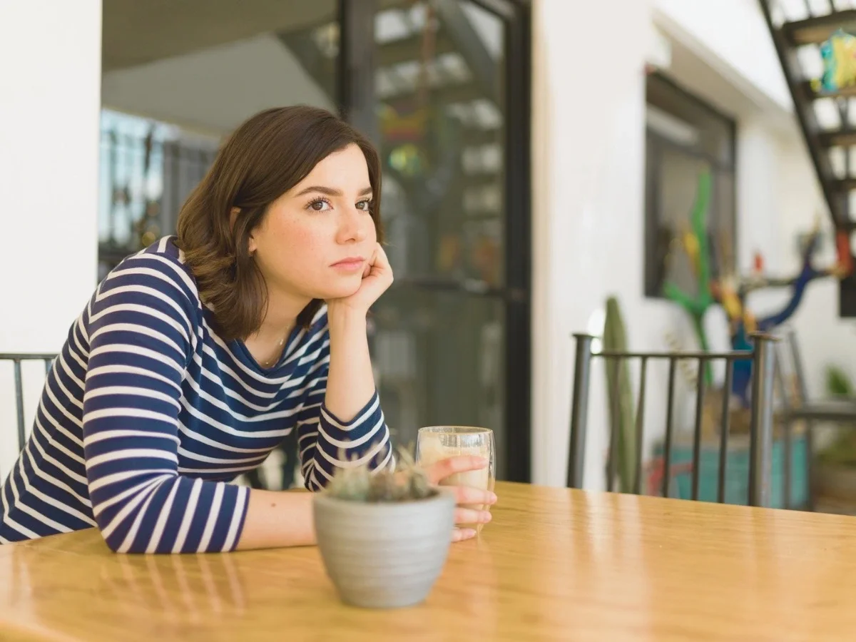 Woman sitting alone at a table holding a drink, looking away thoughtfully — representing the internal experience of emotional suppression in women.