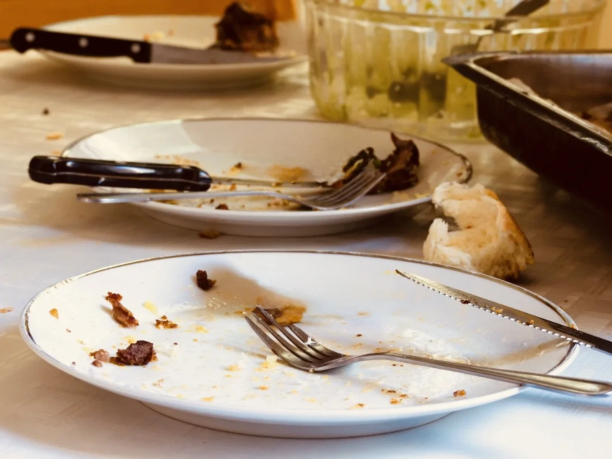 Domestic dining table after a shared meal — two used plates, scattered crumbs, torn bread on a white tablecloth.
