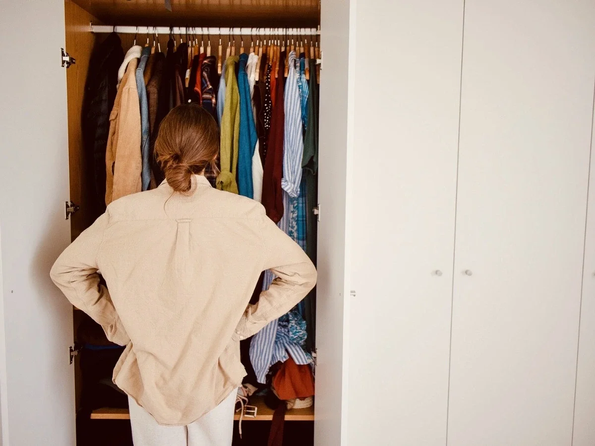 Woman standing with hands on hips facing an open wardrobe full of colorful clothes, representing a moment of reflection during a personal reinvention