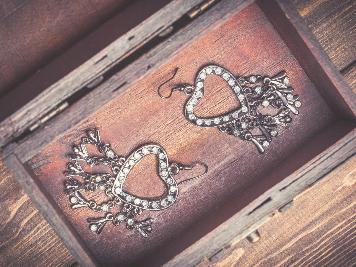 A pair of ornate rhinestone heart-shaped earrings resting unworn inside an open wooden jewelry box — illustrating "What Actually Needs to Change," the section of She The King's Apfel essay on the accessories we buy but rarely wear.