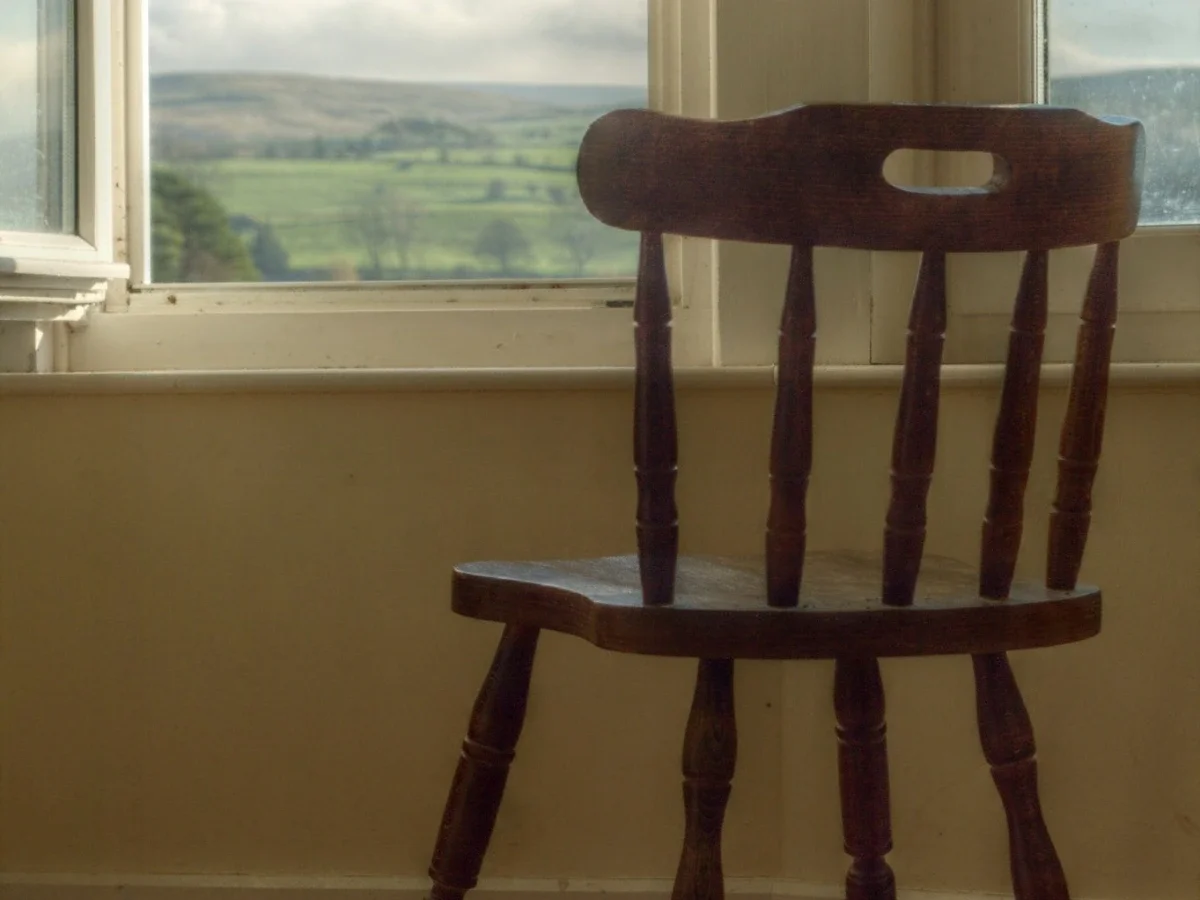 Empty wooden chair positioned before a window overlooking green countryside hills