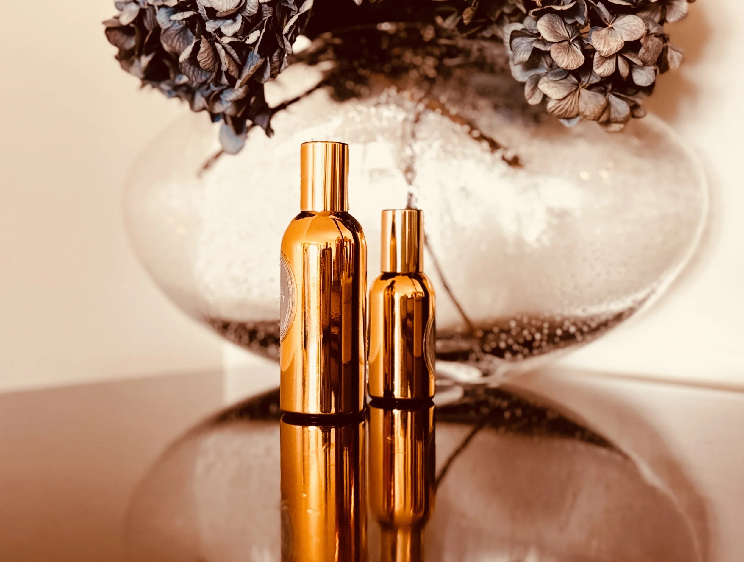Two gold perfume bottles of different sizes on a reflective surface with dried hydrangeas, evoking the idea of fragrance as a sensory autobiography across different life chapters