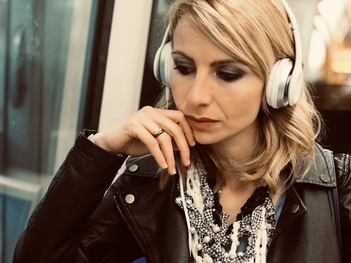 Woman wearing white over-ear headphones sits on a train, looking downward with a distracted expression, chin resting on her hand.