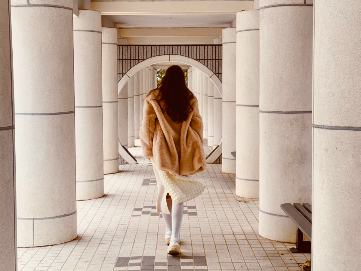 A woman in a fur coat walking alone through a symmetrical white stone colonnade