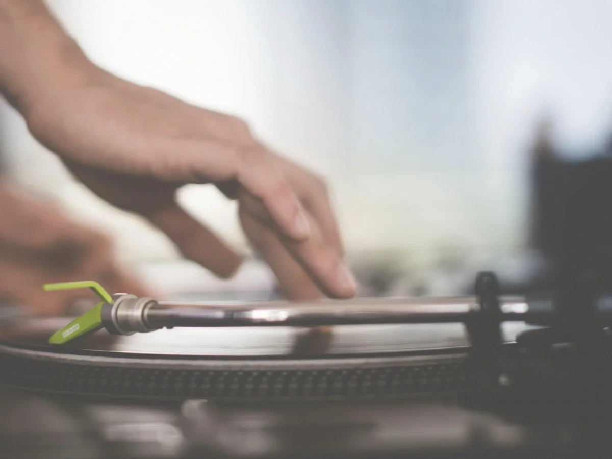 A softly blurred hand reaching toward a spinning turntable with a green cartridge, shot in motion—accompanying "We Gained Almost Everything," on abundance and attention in She The King's music-history essay.