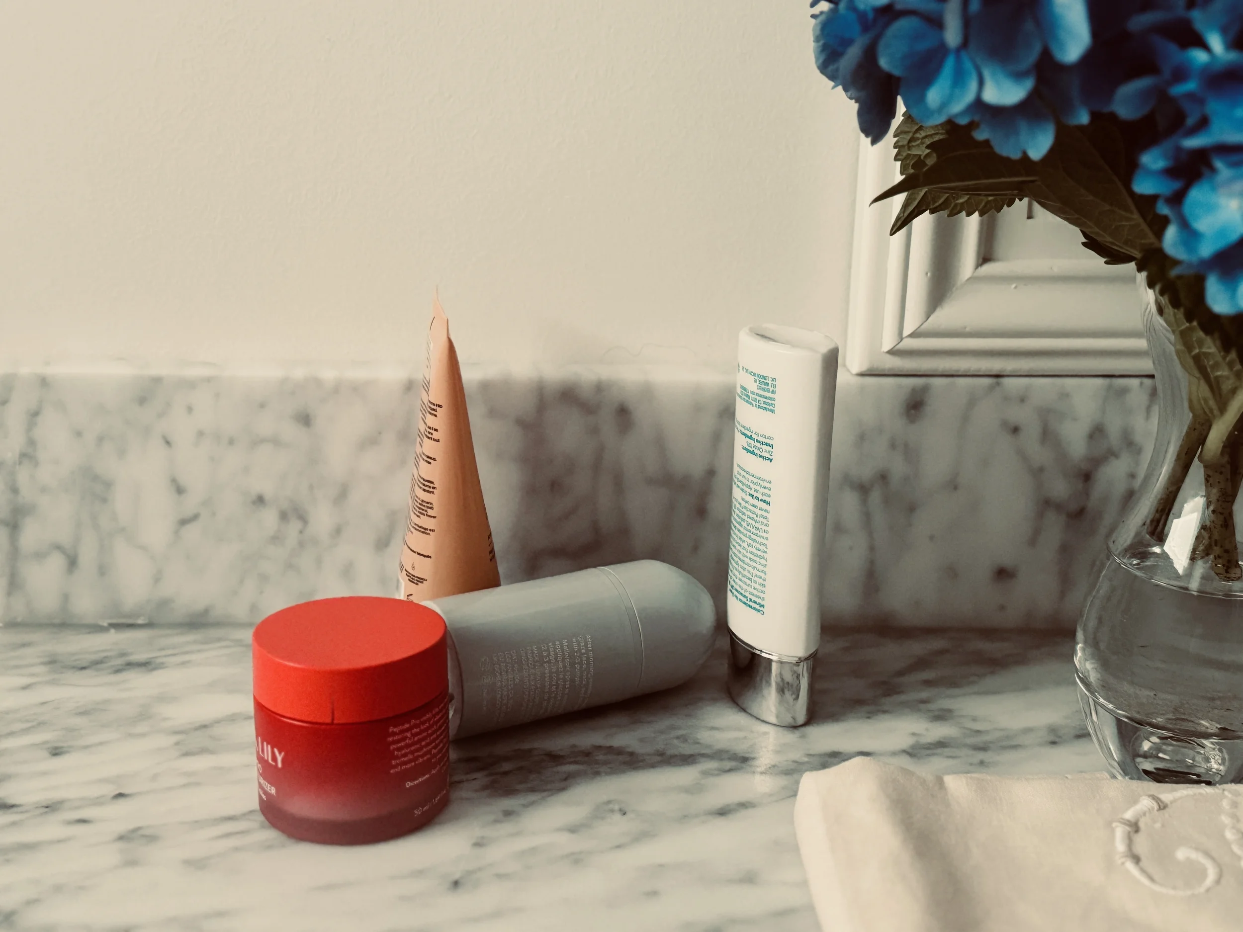 Skincare products arranged on a marble counter beside a vase of blue hydrangeas, soft natural light