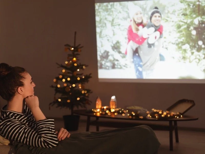 A woman relaxing with warm lights and a Christmas tree while watching a holiday movie, creating a calm and cozy Snuggled Season atmosphere.