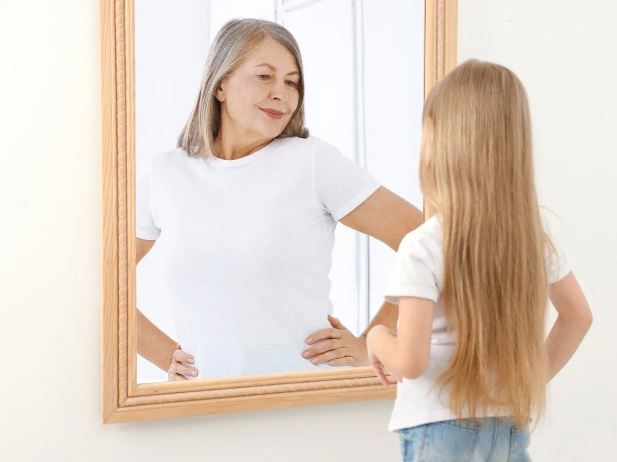 A young girl standing before a mirror, her reflection showing an older woman — a visual metaphor for the inner voice we inherit over time.