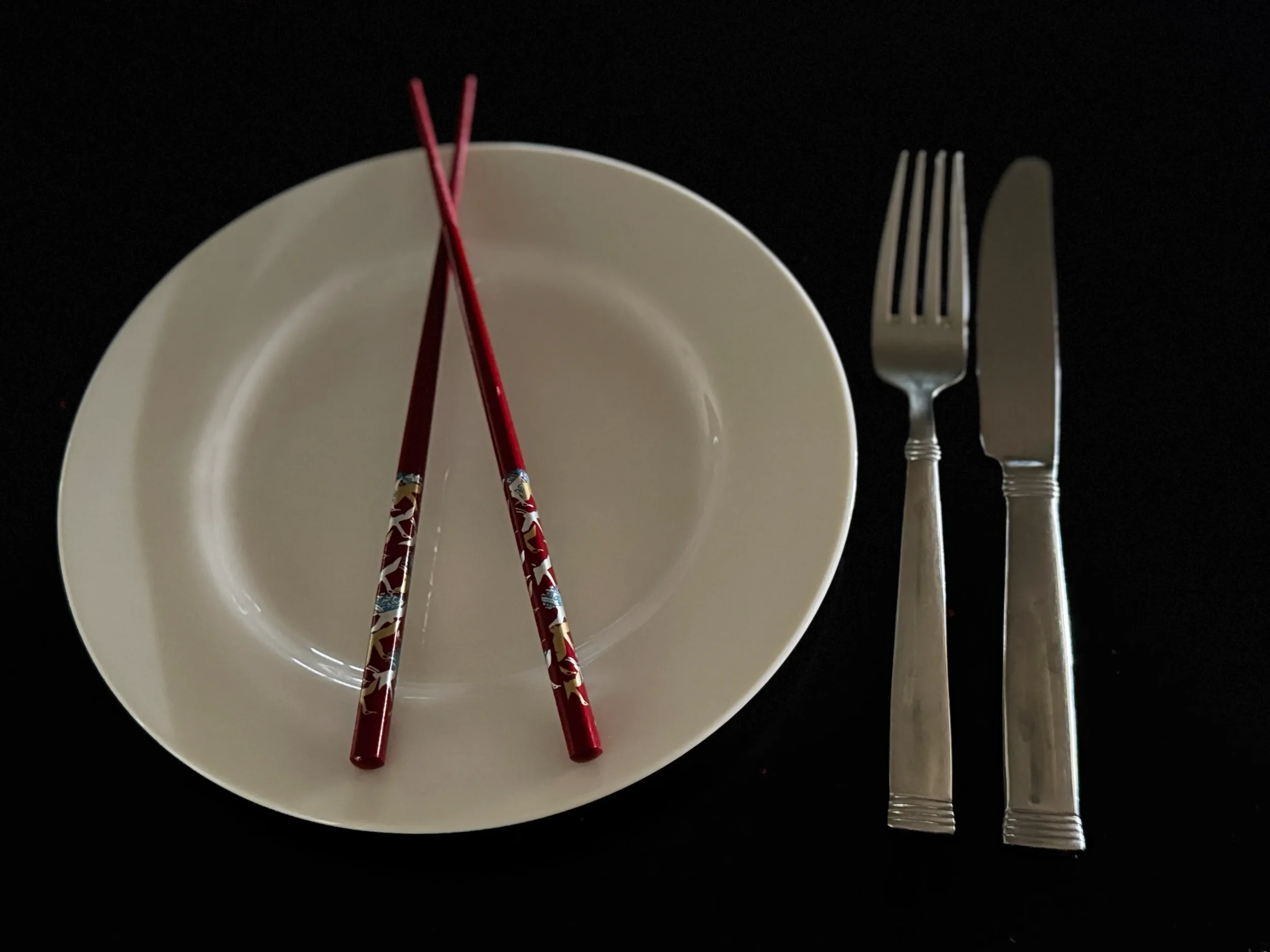 An empty white dinner plate with ornate red lacquer chopsticks resting across it, alongside a Western fork and knife on a dark table, representing the Korean-English cultural duality at the heart of Minbak by Ela Lee