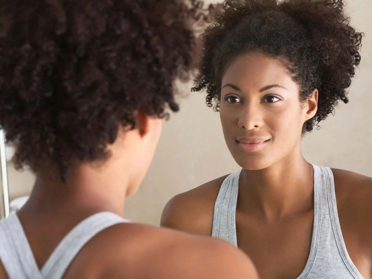 A Black woman looking calmly at her reflection in a mirror, embodying quiet self-awareness and self-compassion