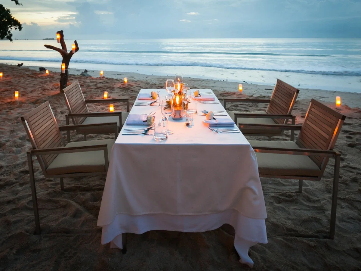 Candlelit beach dinner table set beside the ocean at golden hour with soft coastal lighting and an intimate summer atmosphere.