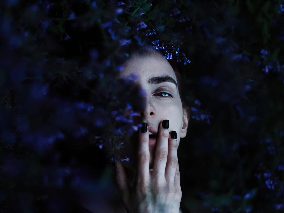 Moody portrait of a woman's face half-hidden behind dark purple flowers and her own hand, one eye visible, evoking guardedness and quiet withholding