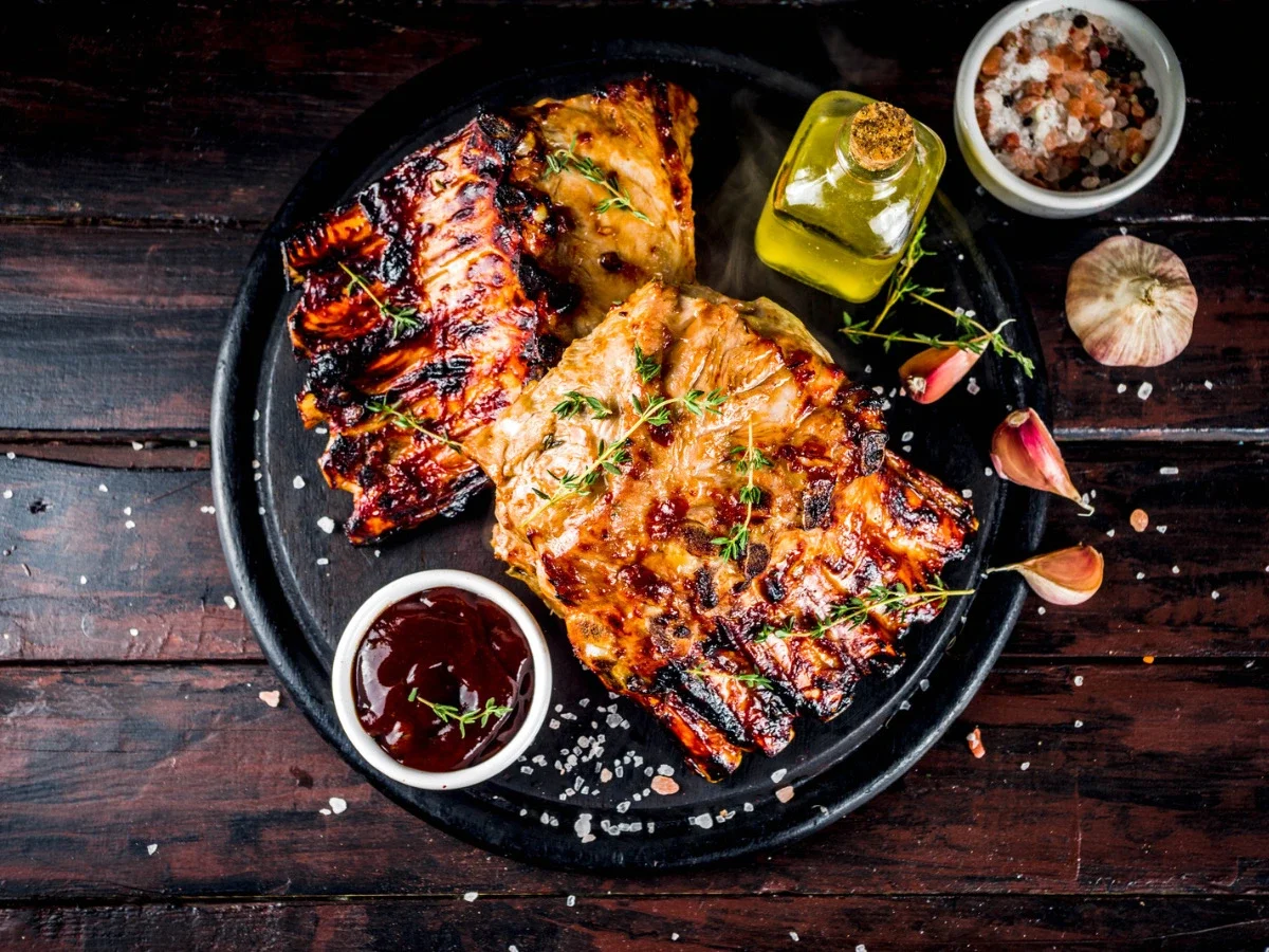 Charred barbecue baby back ribs with thyme and garlic on a dark platter — the Fourth of July pairing for Connor's chapters in The Women
