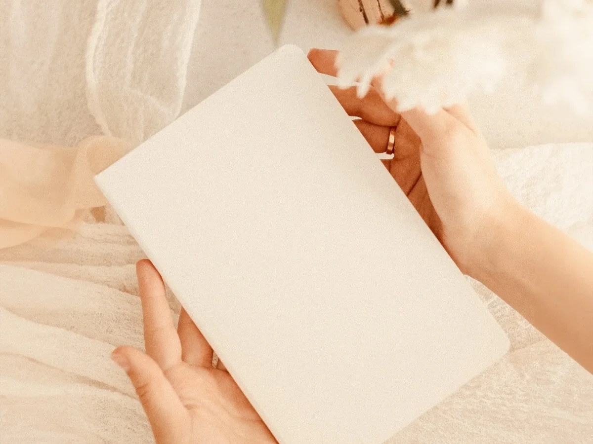 Two hands hold open a blank cream-coloured journal page in soft warm light against white linen. A gold ring is visible. The page is empty — held but not yet written in.