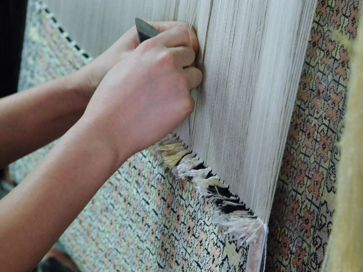 A close-up of two hands working a vertical loom, a metal tool pulling a single thread through densely strung warp threads. The intricate pattern of the textile in progress is visible below — the result of sustained technical mastery.