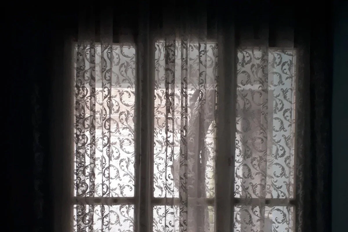 Soft light through lace curtains in a quiet home, symbolizing inherited silence and post-war displacement in The Safekeep.