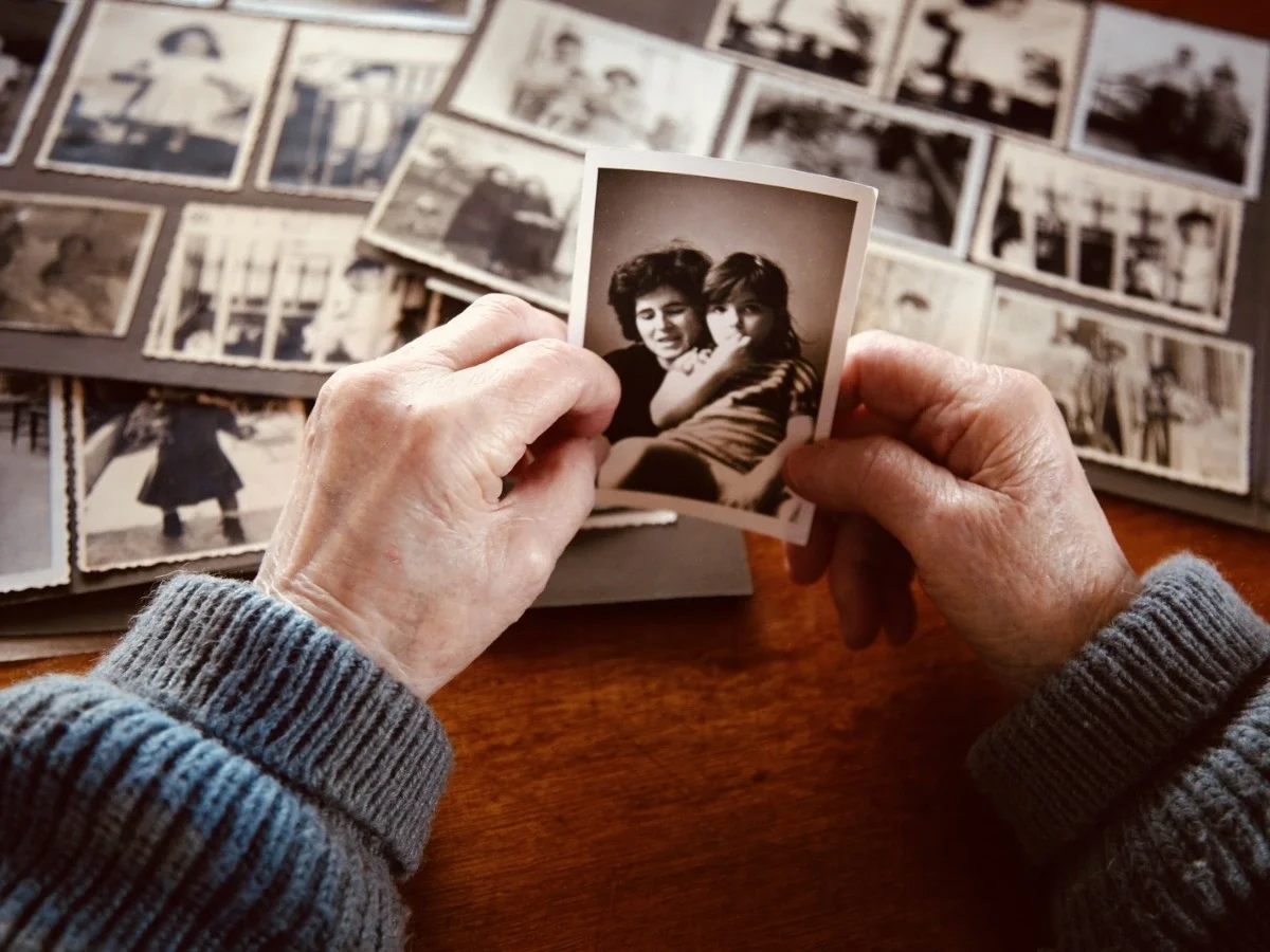 A pair of elderly hands in a grey knit sweater hold a small black and white photograph of two people against a warm wooden surface, surrounded by other vintage photographs.